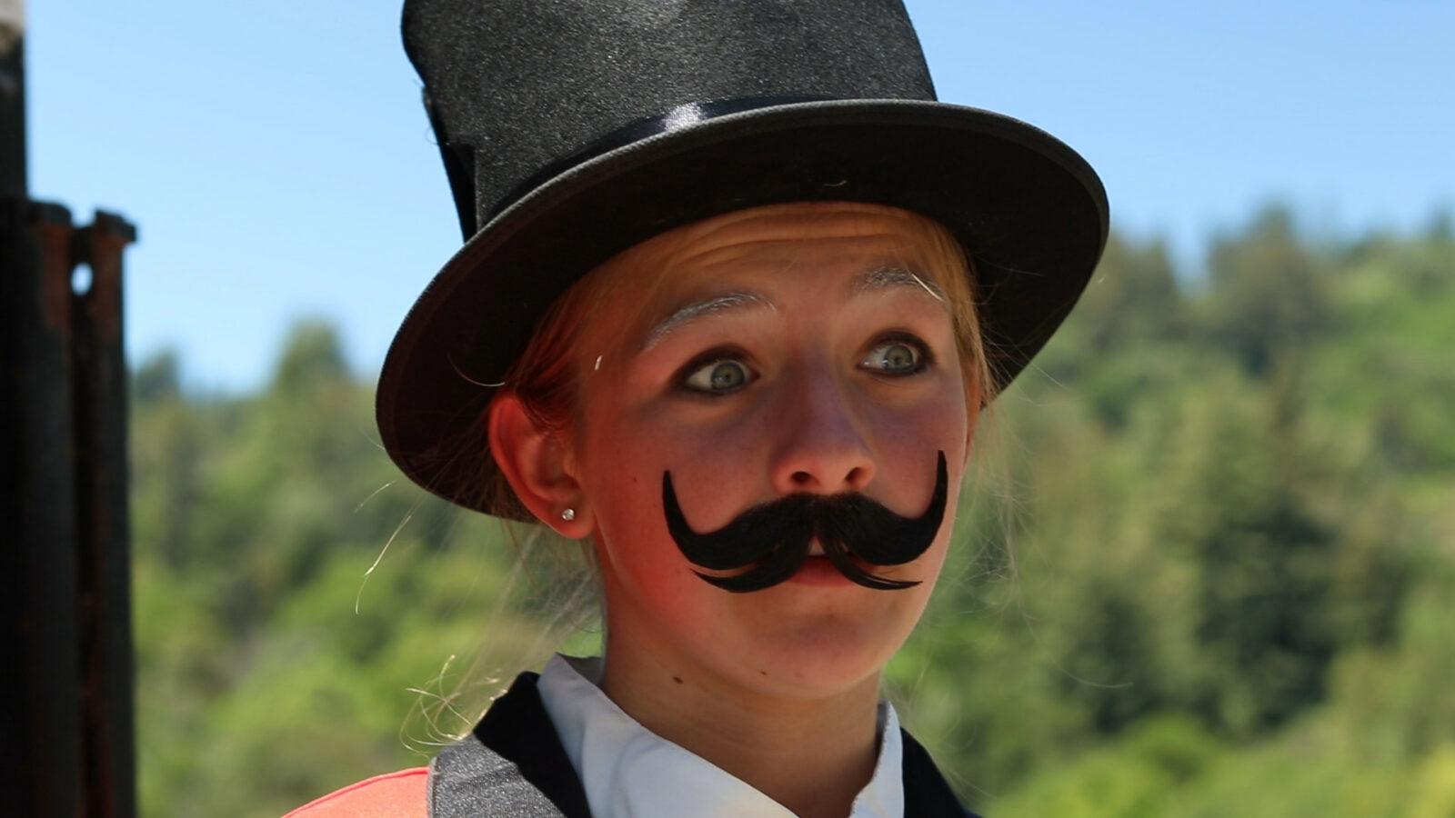 A girl with a top hat and mustache on.